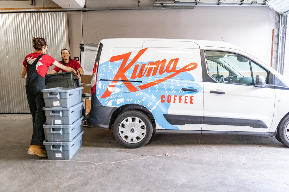 Two women wearing red bear t-shirts with a logo for Kuma Coffee smiling and lifting bins into the back of a van with the Kuma Coffee logo over a large blue bear on the side