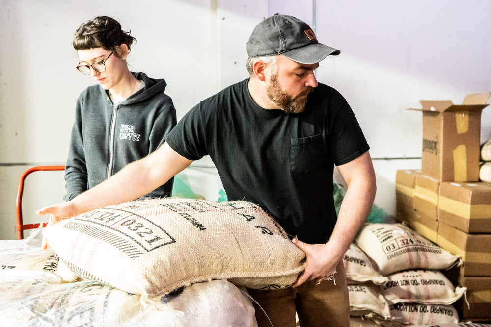Man and woman wearing Kuma Coffee sweatshirt hard at work lifting burlap bags filled with Colombian coffee