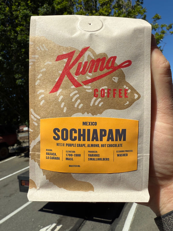 A hand holding a bag of Kuma Coffee Sochiapam chocolate coffee. The bag is off-white with a brown bear graphic, a red Kuma logo, and a yellow label indicating it is a mixed fruit chocolate with almonds and chocolate chips from Mexico.